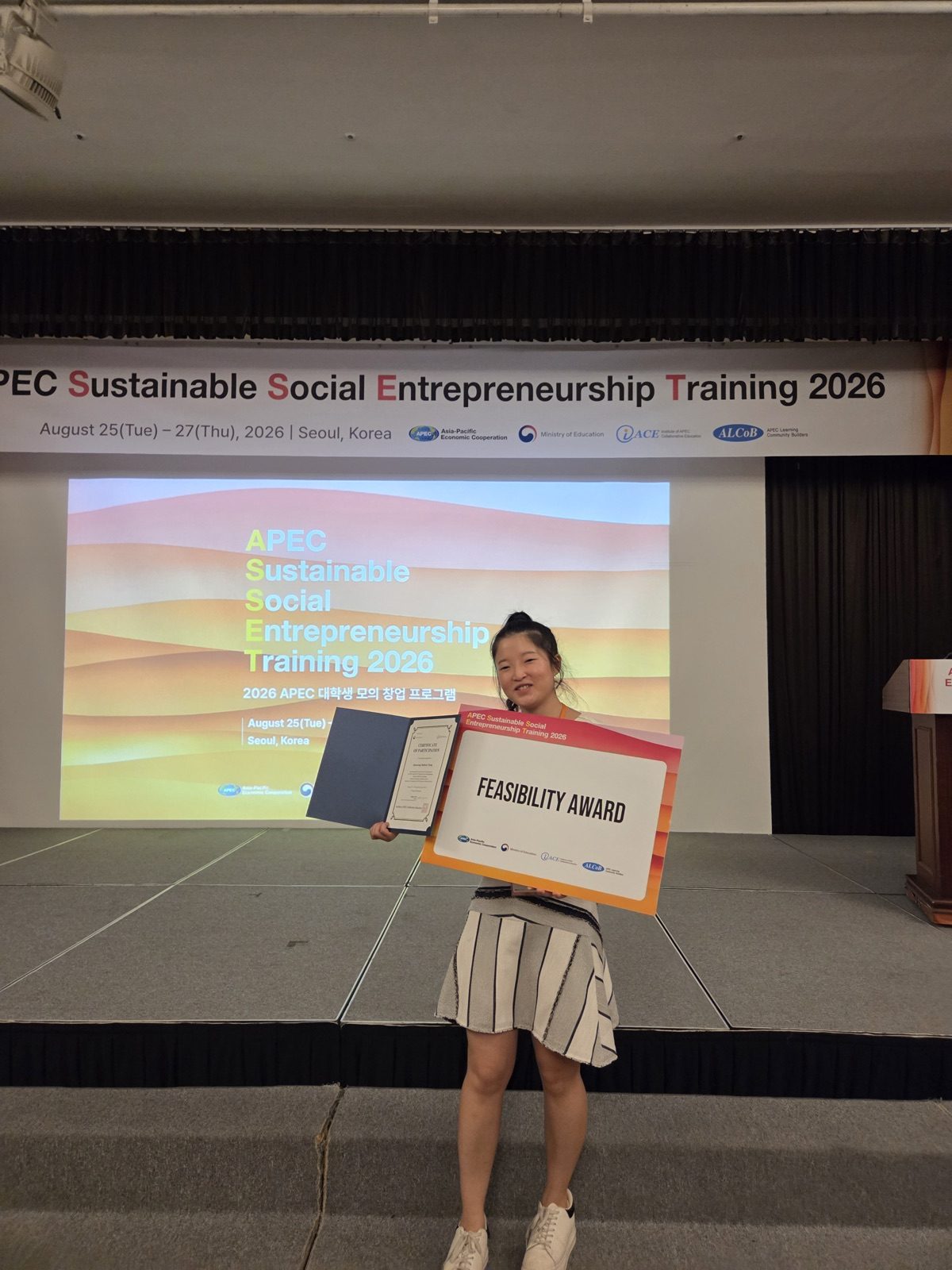 APEC 프로그램 현수막이 걸린 무대 앞에서 정진서랑이 참가 증서와 ‘FEASIBILITY AWARD’ 판을 들고 웃고 있다.